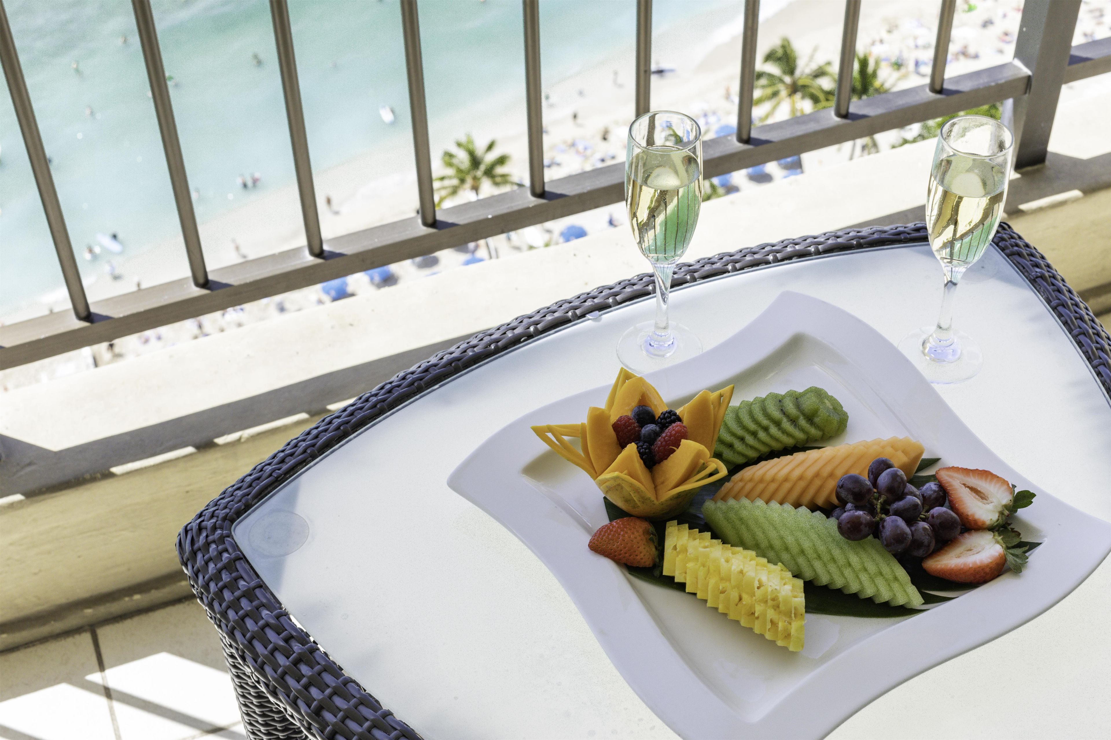 Uma mesa com uma cesta de frutas e duas taças de vinho branco numa varanda com vista para a praia.