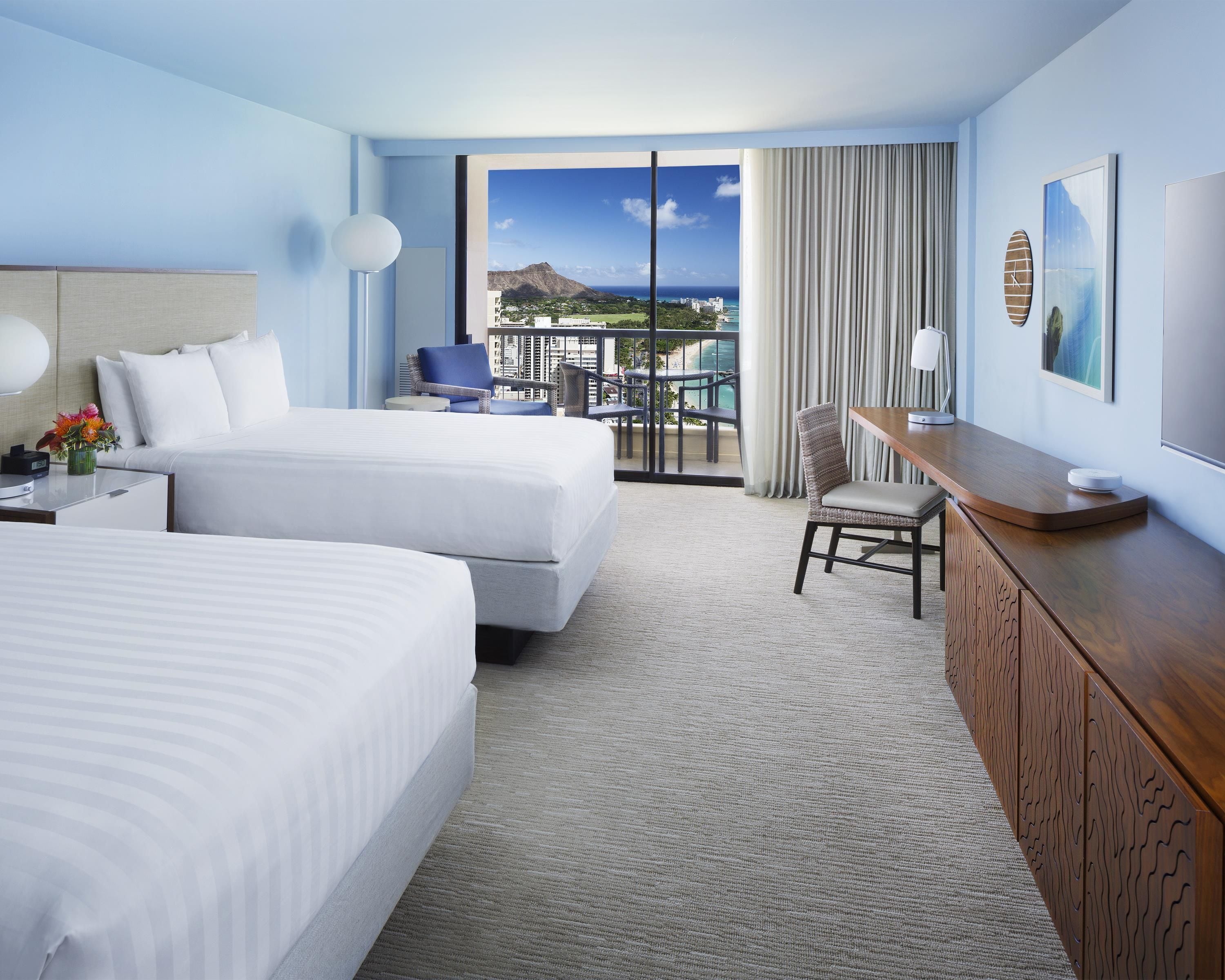 A hotel room with two beds, a desk, a chair, a TV, and a view of the ocean and buildings.