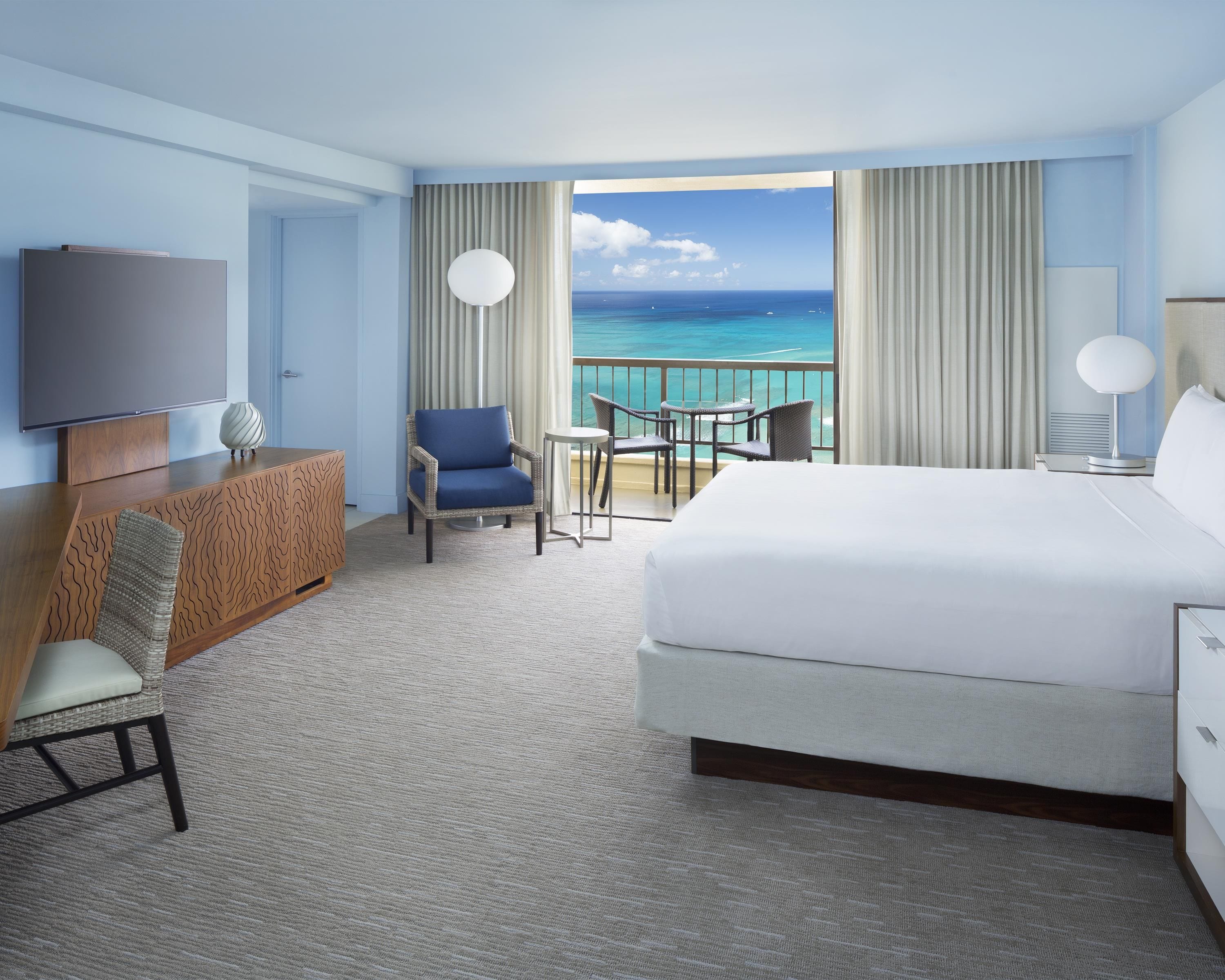 Habitación de hotel con una cama grande, televisión, escritorio con silla, una mesita con sillas y balcón con vista al océano.