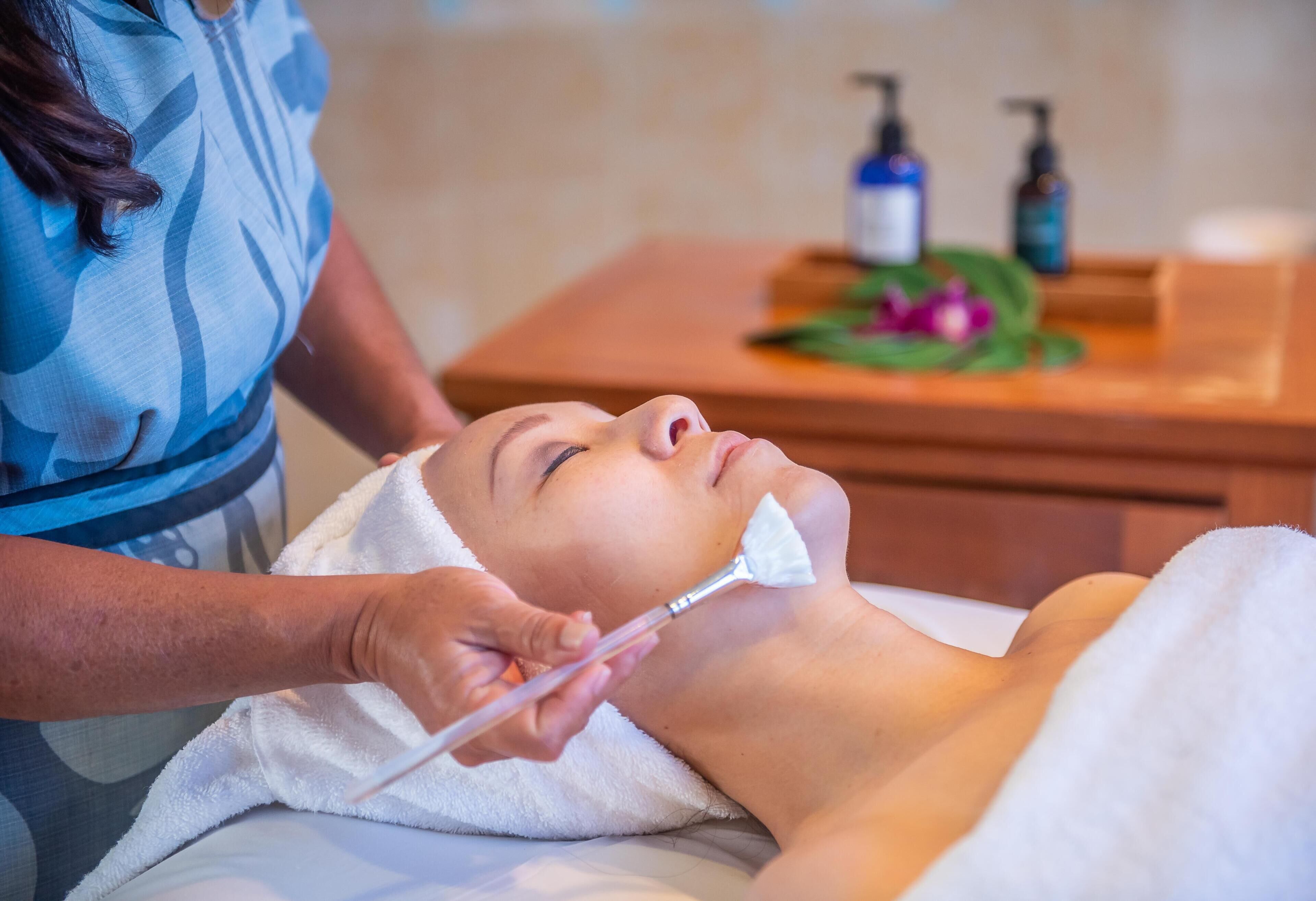 A person receiving a facial treatment in a room with a wooden side table, a towel, and a bottle of liquid soap.