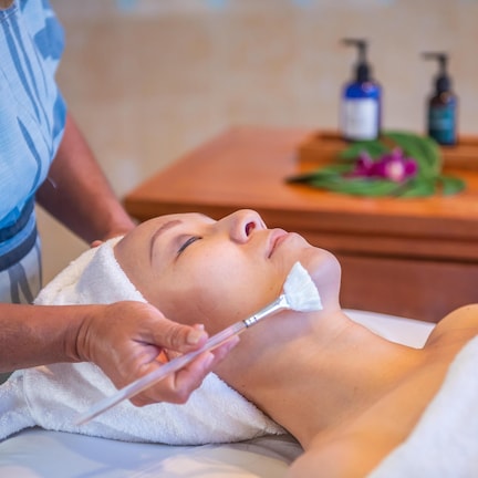 A person receiving a facial treatment in a room with a wooden side table, a towel, and a bottle of liquid soap.