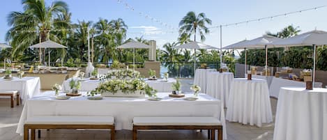 An outdoor dining area with white tables and benches, string lights, and palm trees in the background.