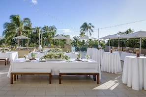 An outdoor dining area with white tables and benches, string lights, and palm trees in the background.