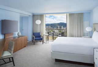 A hotel room with a bed, a TV, a desk, a chair, a balcony with a view of a cityscape.