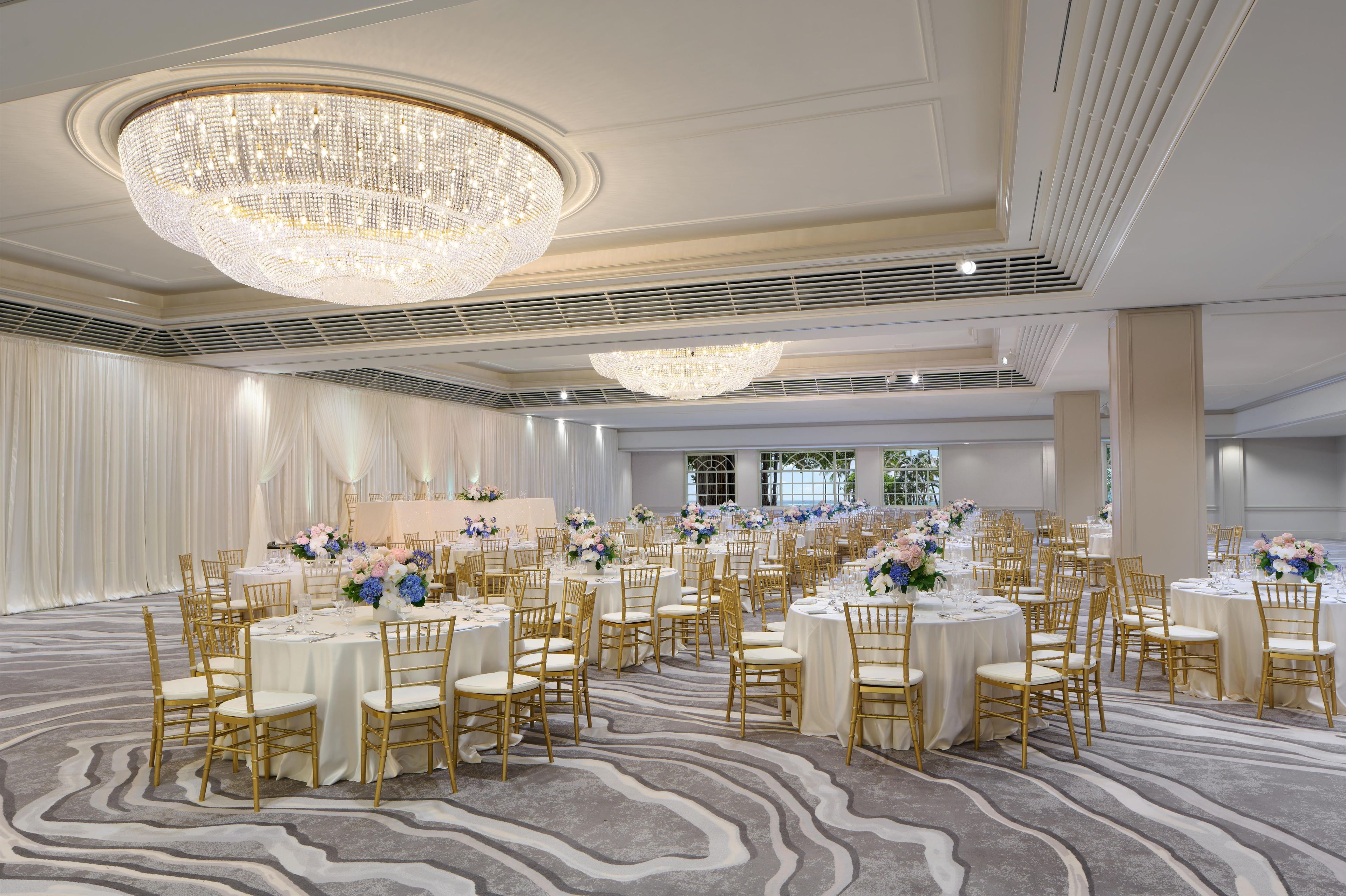 A banquet hall with round tables, gold chairs, and a large chandelier.