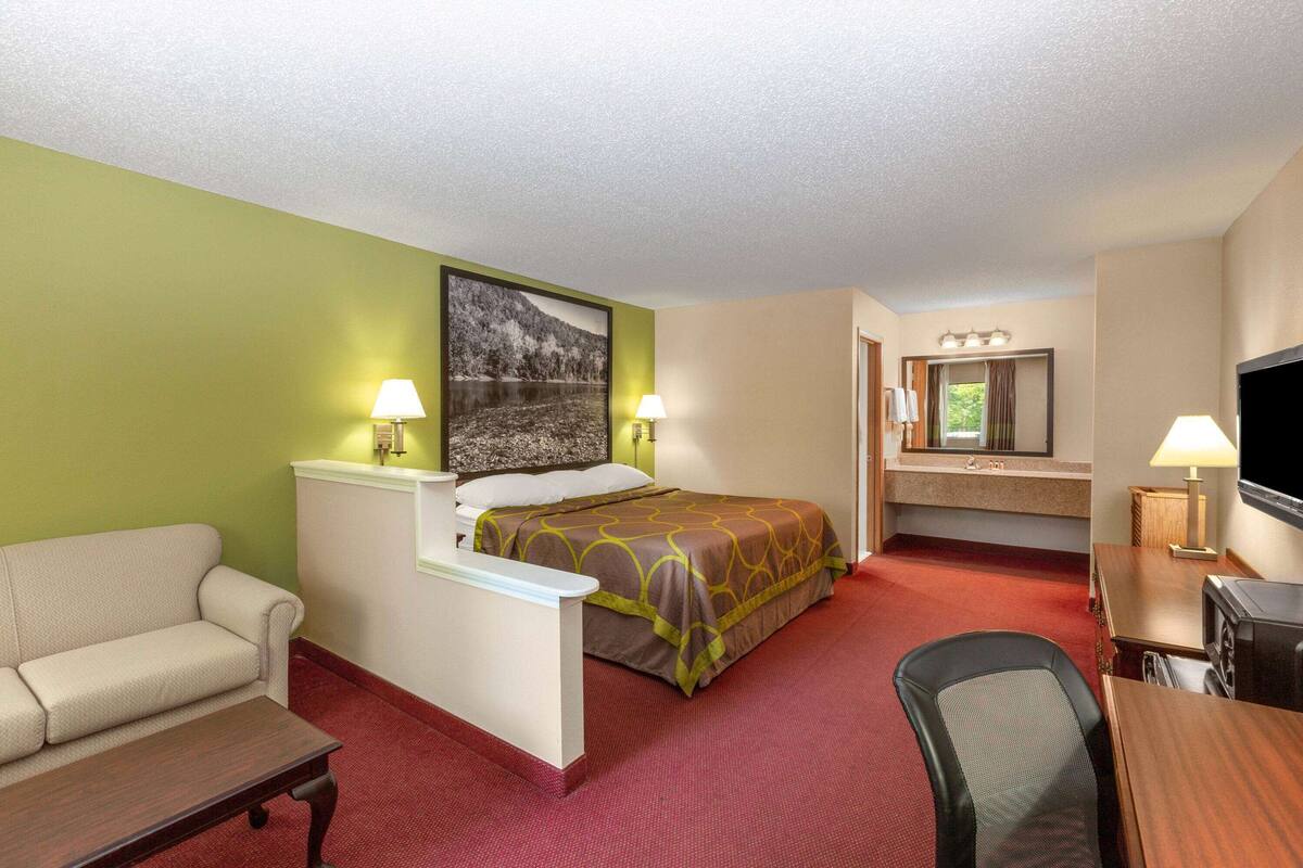 Habitación de hotel con cama, sofá, escritorio, televisor y baño con espejo y lavamanos.