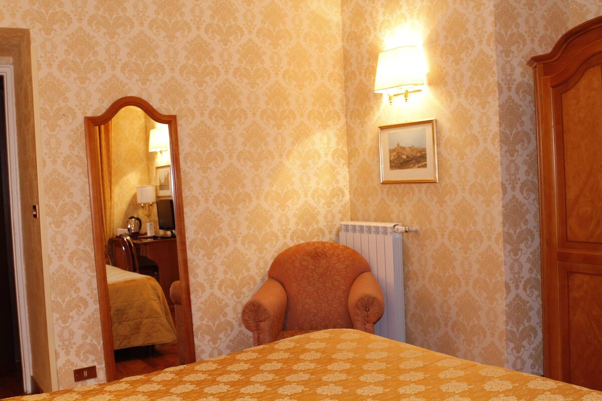 Habitación de hotel con cama, una silla, un espejo, un radiador y un cuadro colgado en la pared.
