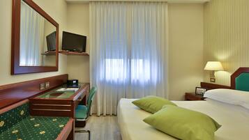 Een hotelkamer met een bed, een bureau met stoel, een televisie, een raam met gordijnen en een nachtkastje met lamp.