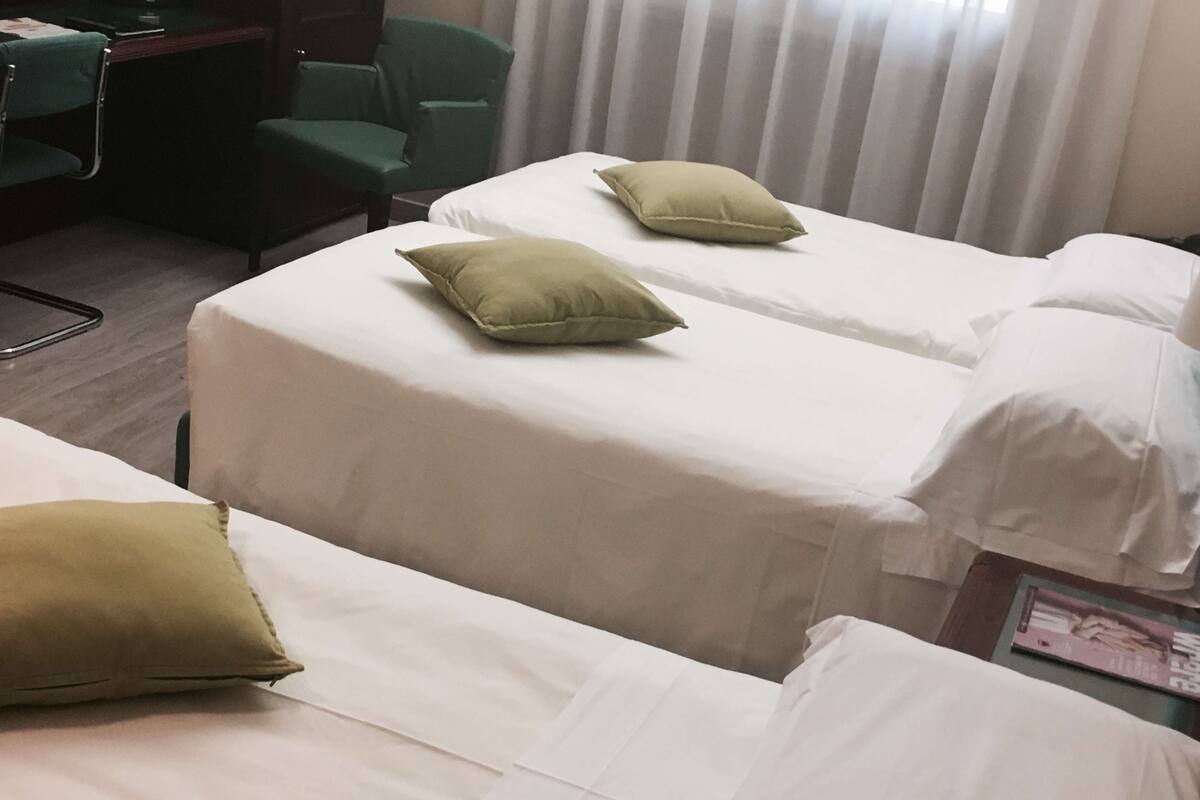 A hotel room with two single beds, each with a pillow and a white bedspread. There is a desk with a chair and a window with blinds.