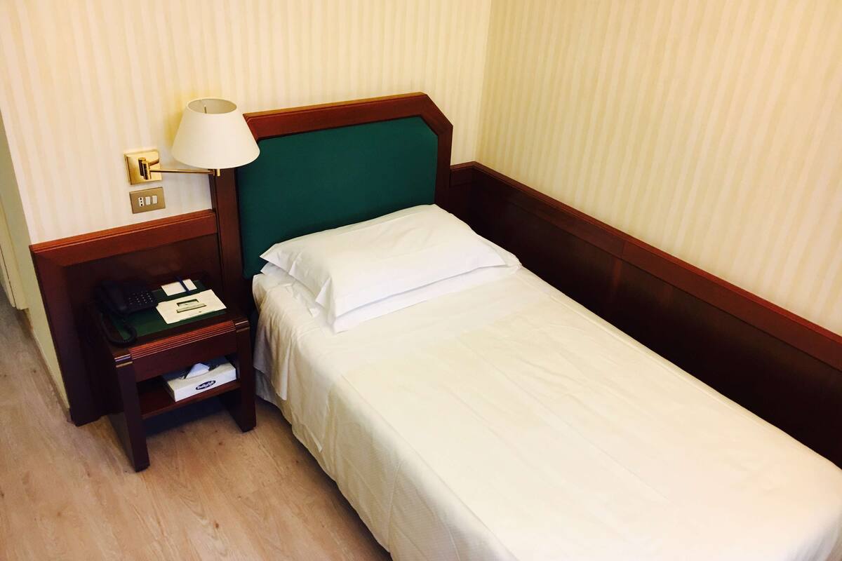 A single bed with a green headboard, a bedside table with a lamp, a phone, and a notepad.