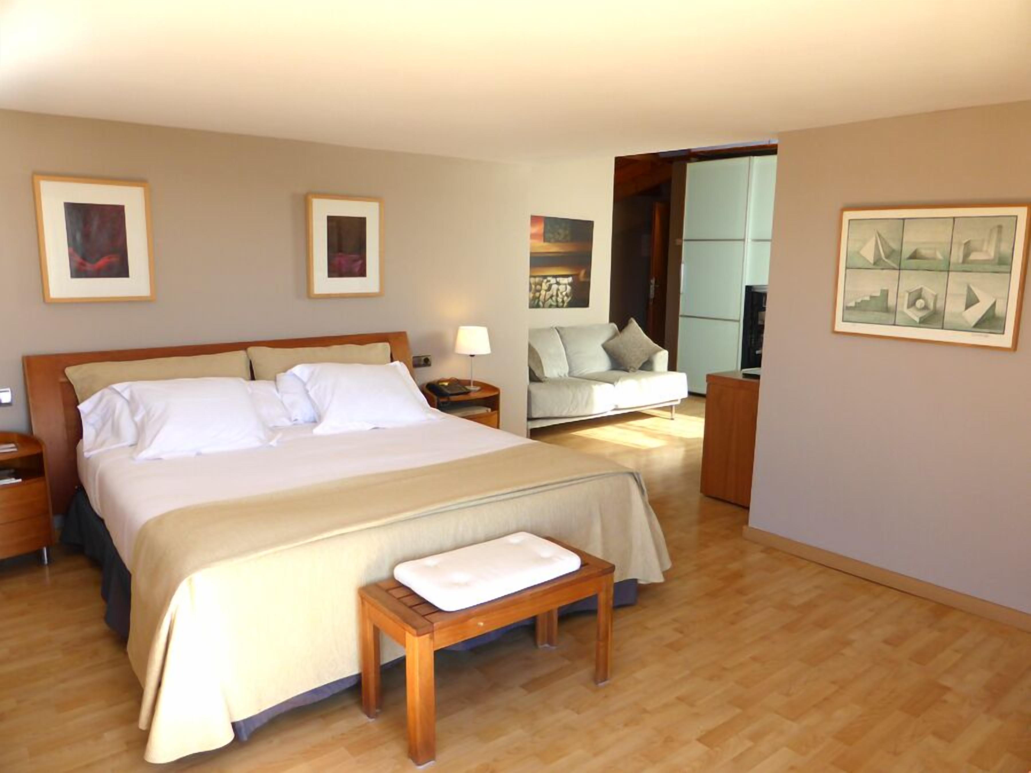 Habitación de hotel con una cama grande, una mesita de noche, un banco, un sofá y una pequeña mesa. Hay cuadros enmarcados en las paredes.
