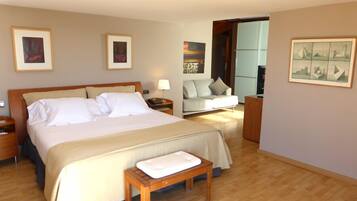 Habitación de hotel con una cama grande, una mesita de noche, un banco, un sofá y una pequeña mesa. Hay cuadros enmarcados en las paredes.