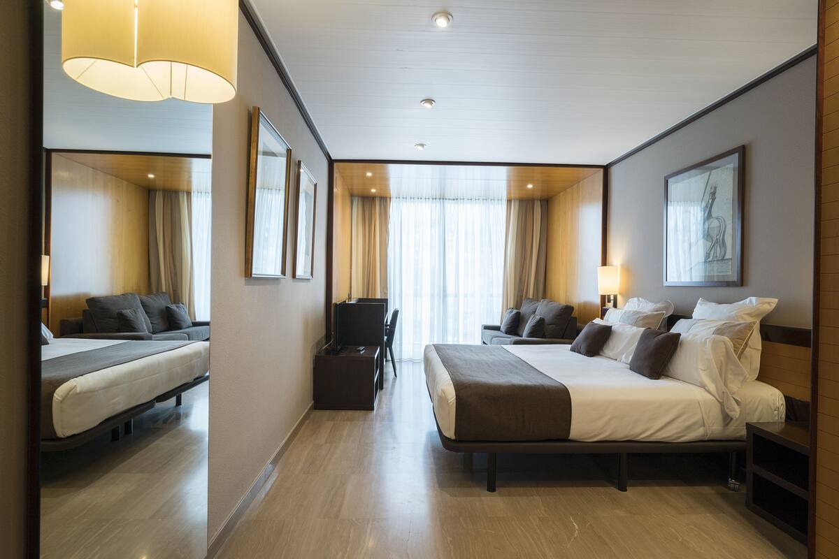 A hotel room with two beds, a desk, a TV, and a large window with curtains.
