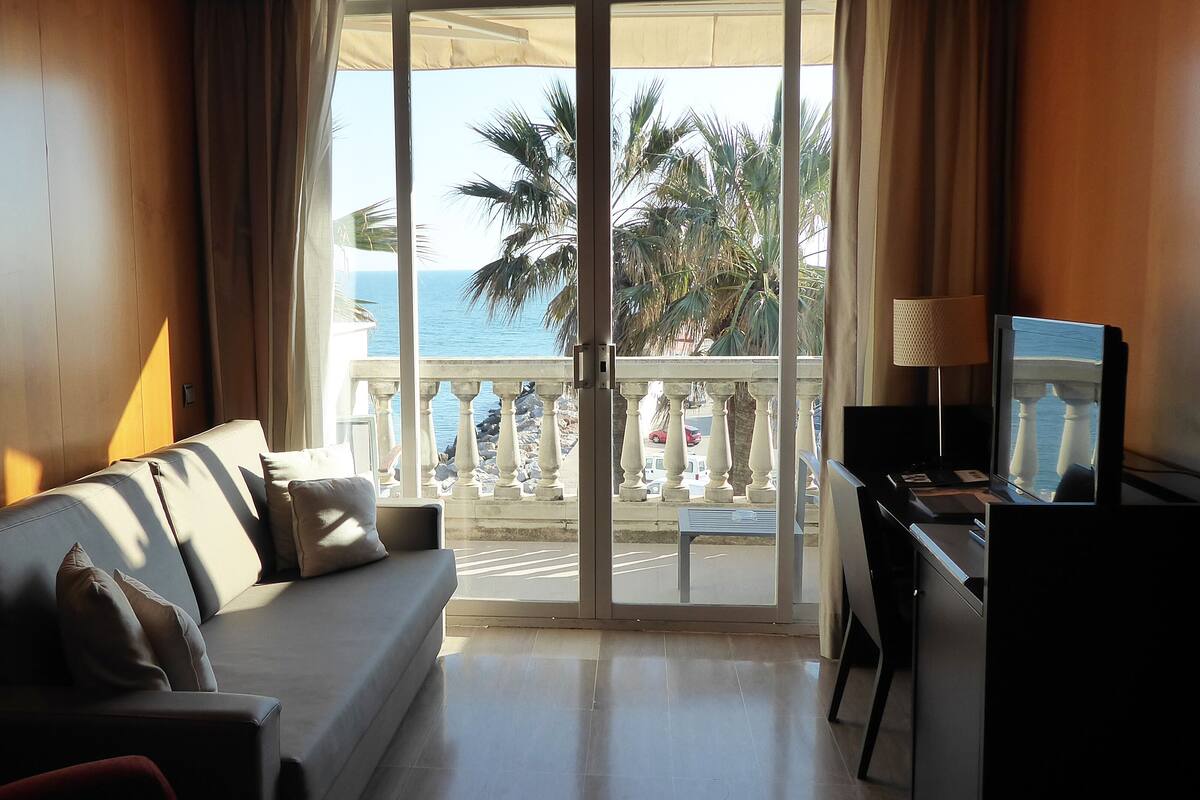A hotel room with a sofa, a TV, and a view of the sea through a balcony.