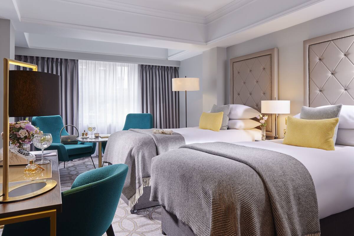 A hotel room with two beds, a desk with a lamp, a chair, and a large window with curtains.