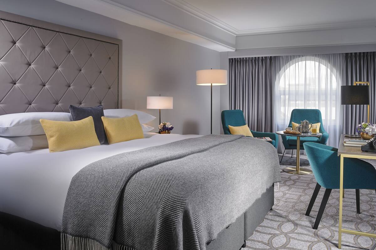 A hotel room with a tufted headboard, a large bed with multiple pillows, a desk with a lamp, and a chair.