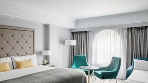 A hotel room with a tufted headboard, a bed with a grey and white bedspread, two teal armchairs, a small round table, and a large window with grey curtains. - The Davenport (Dublin)