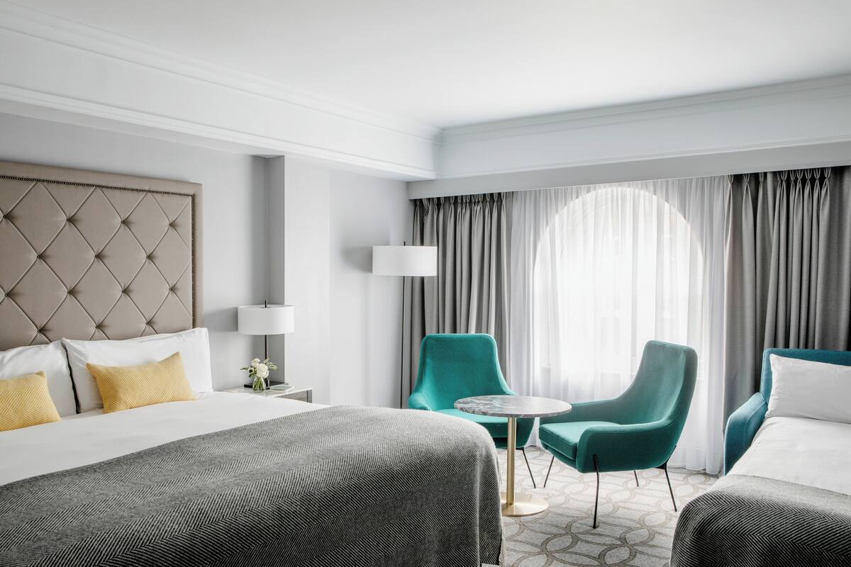 A hotel room with a tufted headboard, a bed with a grey and white bedspread, two teal armchairs, a small round table, and a large window with grey curtains.