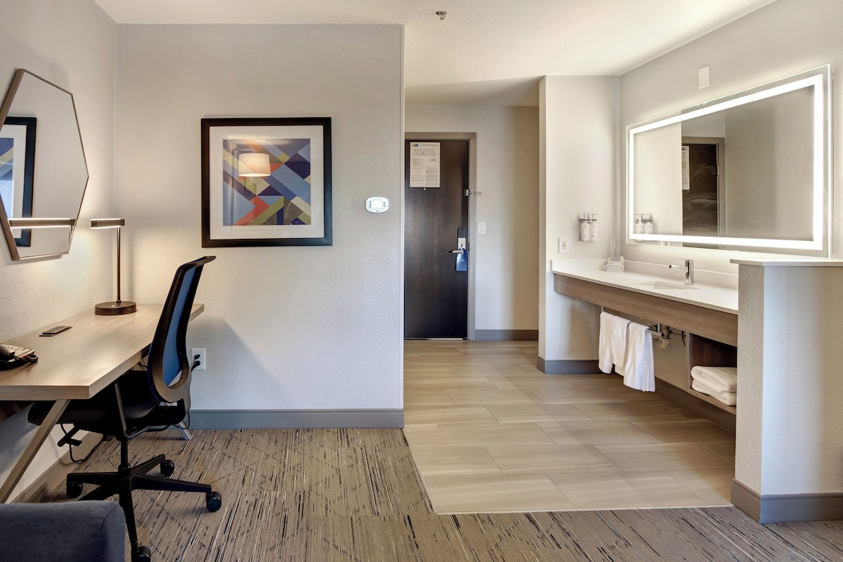 Quarto de hotel com escrivaninha, cadeira, espelho grande e pintura abstrata emoldurada.