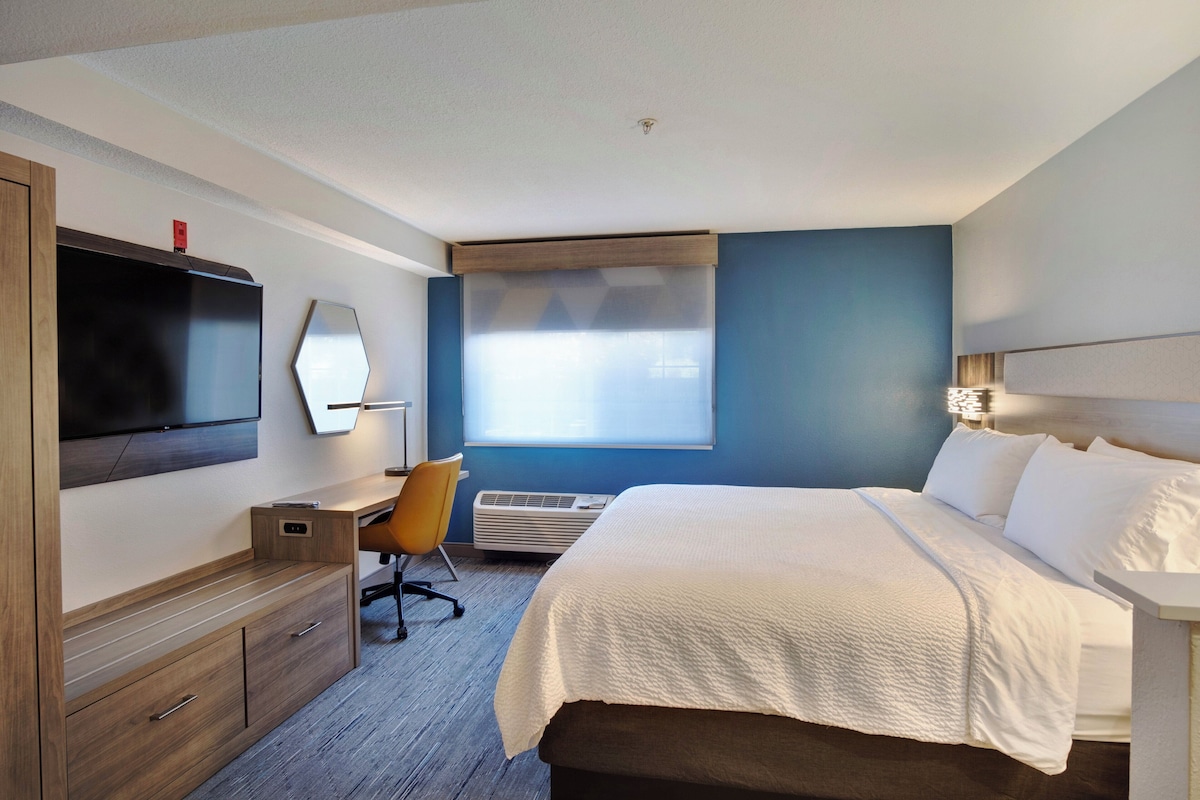 Quarto de hotel com cama, escrivaninha, cadeira, televisão e janela com persianas.