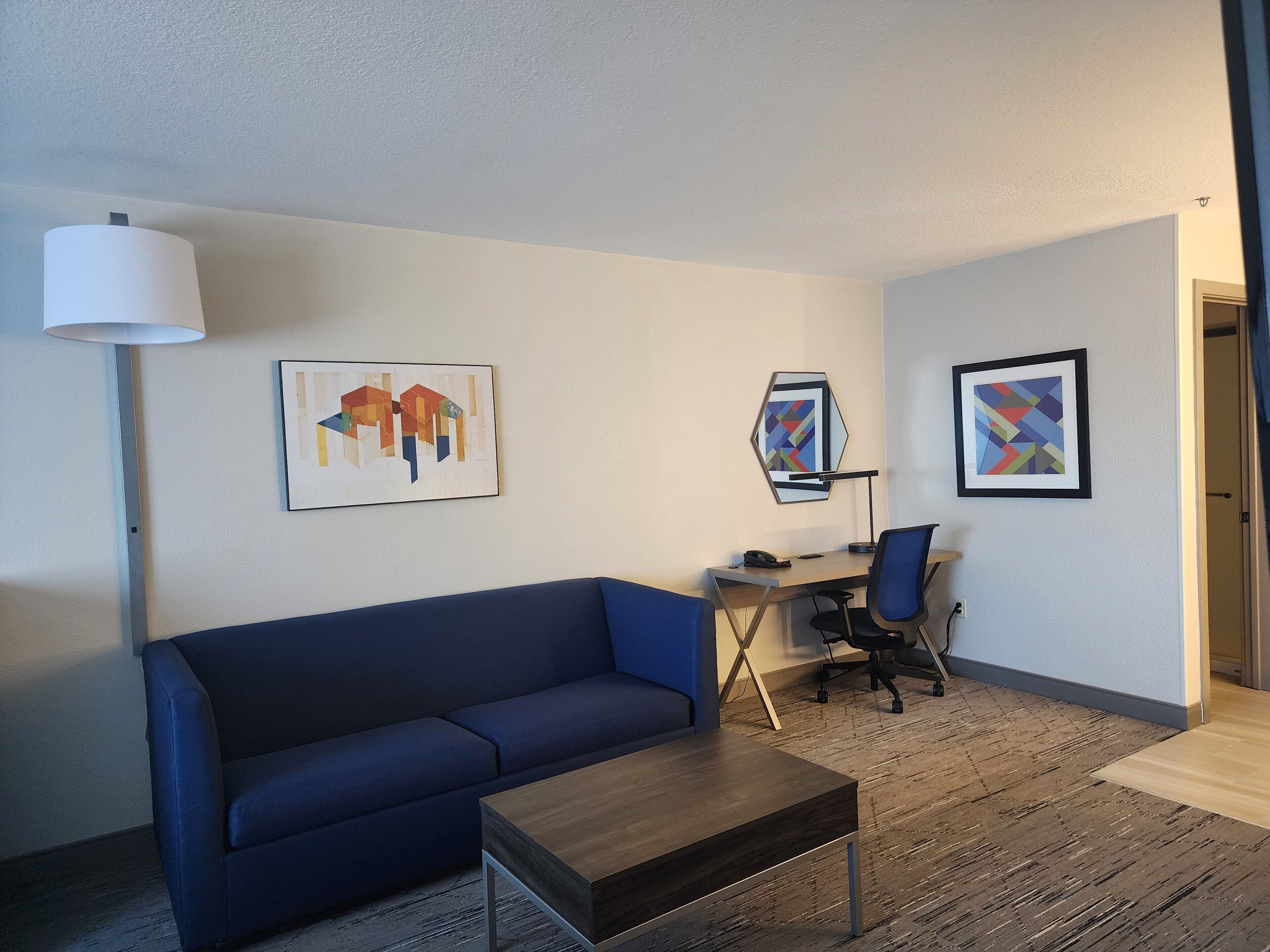 Une chambre d’hôtel moderne avec un canapé bleu, une table basse en bois, un bureau avec une chaise et deux œuvres d’art encadrées accrochées au mur.