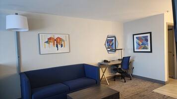 Une chambre d’hôtel moderne avec un canapé bleu, une table basse en bois, un bureau avec une chaise et deux œuvres d’art encadrées accrochées au mur.
