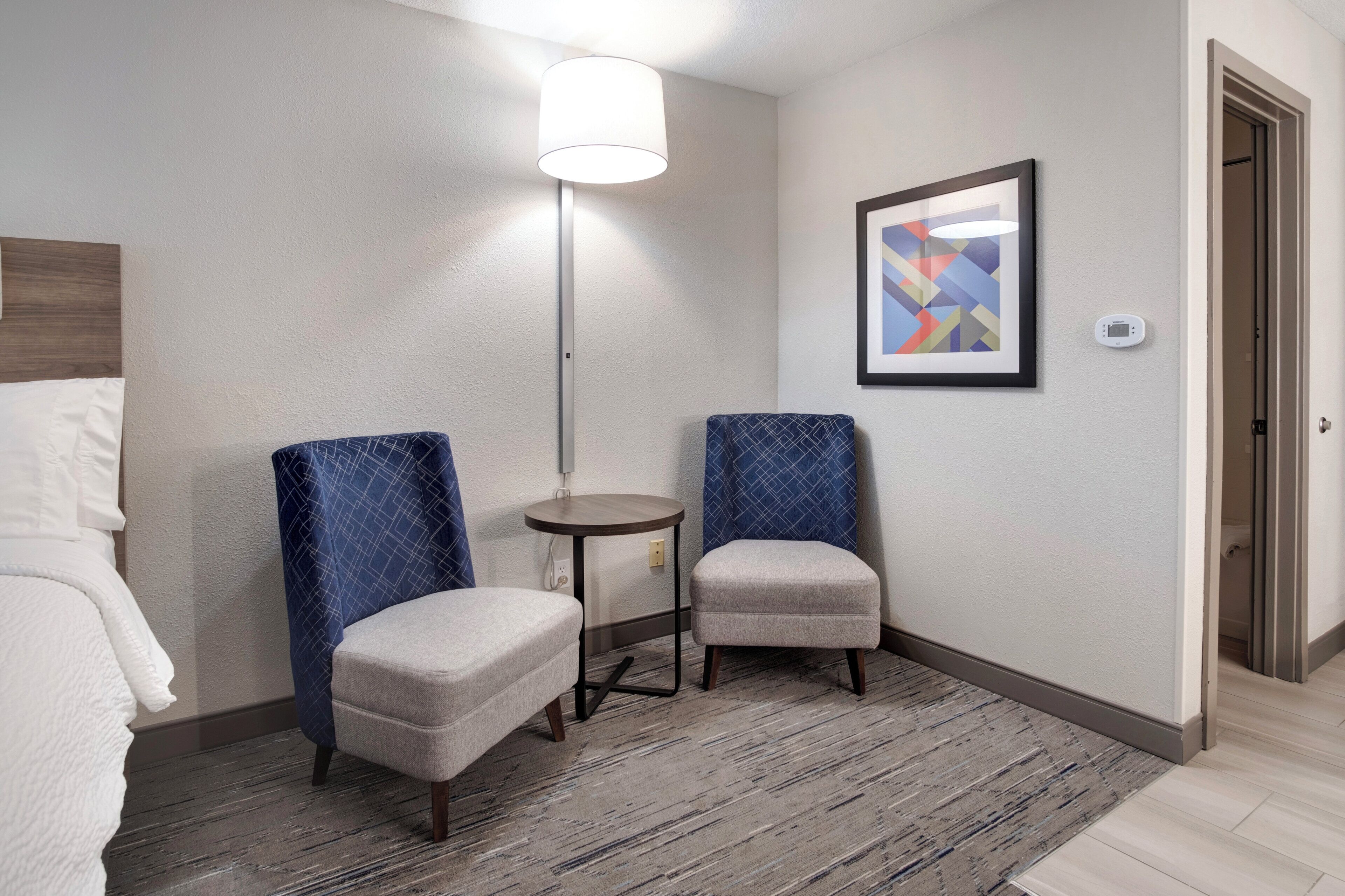 A hotel room with two armchairs, a small round table, a lamp, a framed abstract painting, and a thermostat on the wall.