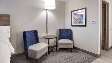 A hotel room with two armchairs, a small round table, a lamp, a framed abstract painting, and a thermostat on the wall.