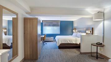 A hotel room with two beds, a desk, a chair, a mirror, a lamp, and a view of the outside.
