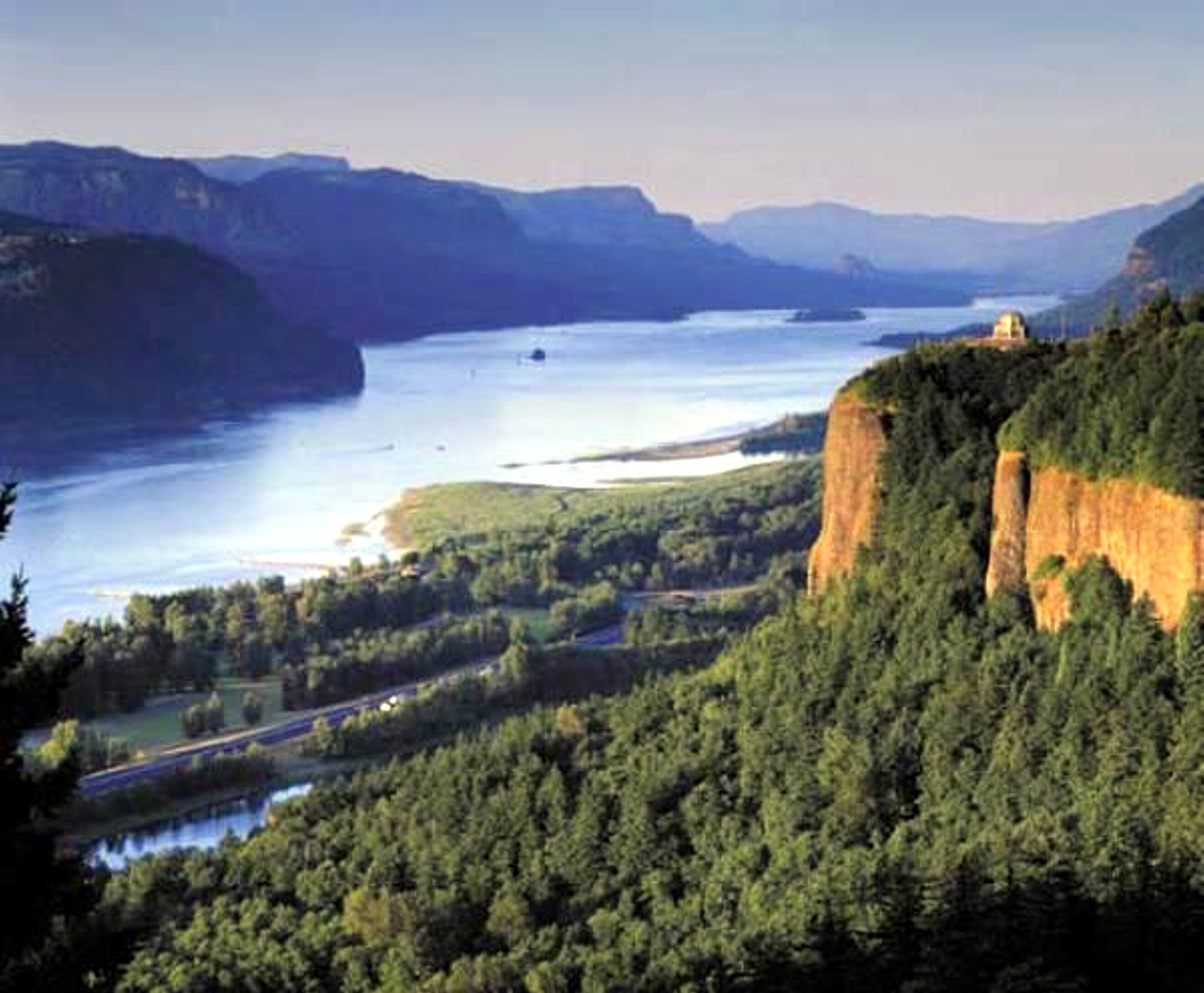 A mountainous landscape with a river, dense forest, and a cliff with a structure on top.
