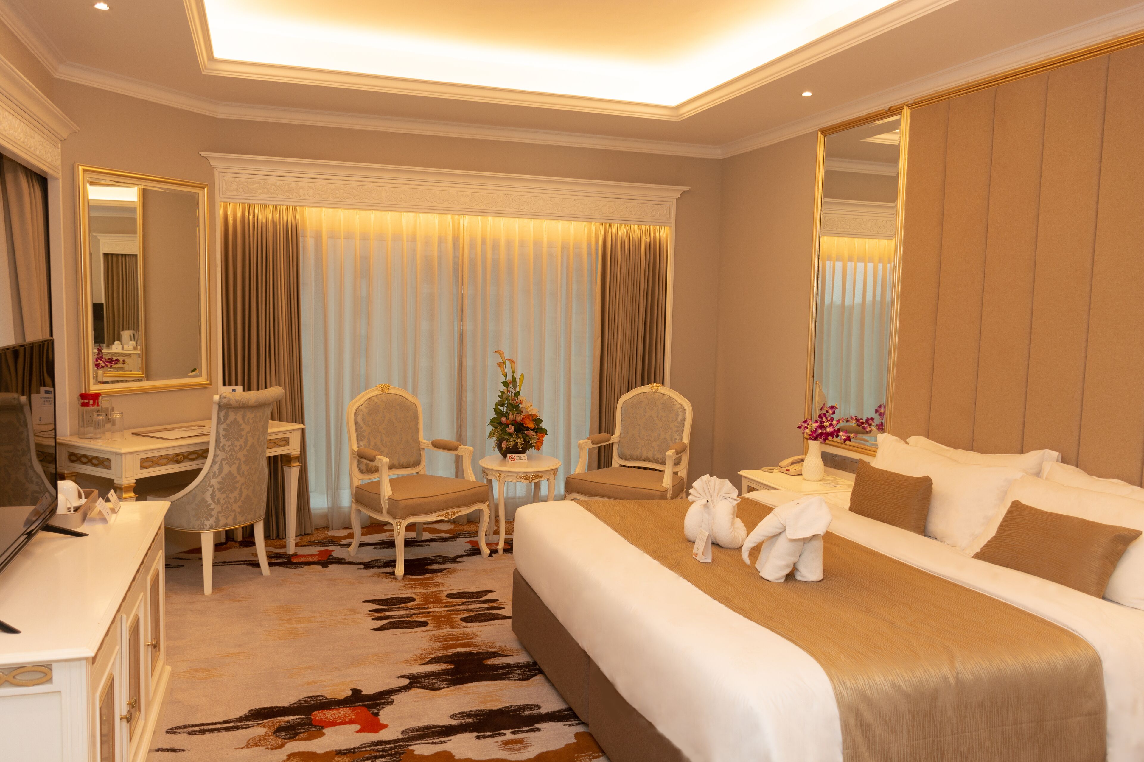 A hotel room with a large bed, two armchairs, a small table with a vase of flowers, a desk with a mirror, and a television.