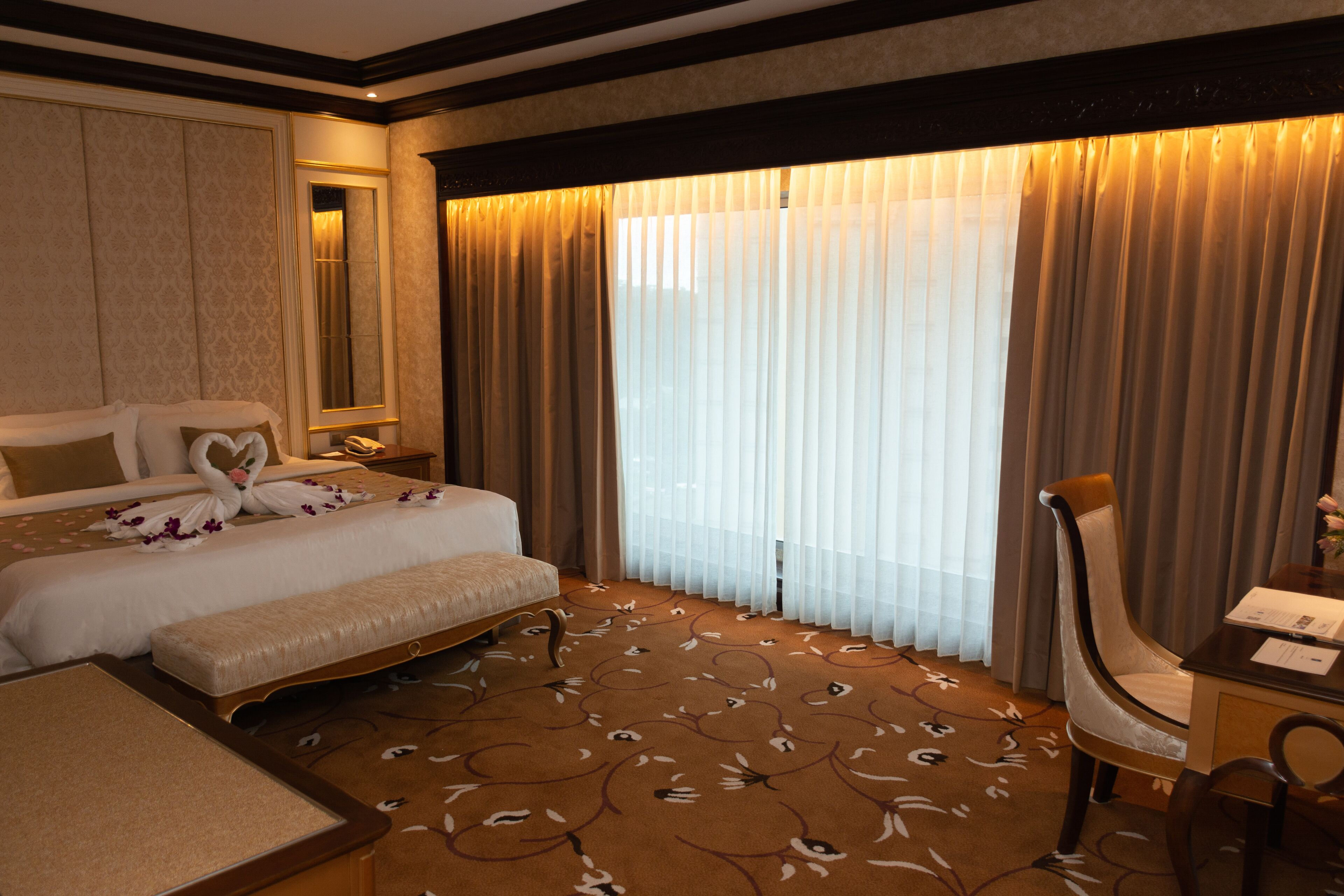 A hotel room with a large bed, a desk, and a chair. There is a window with curtains, a mirror, and a carpet with a floral pattern.