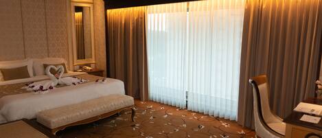 A hotel room with a large bed, a desk, and a chair. There is a window with curtains, a mirror, and a carpet with a floral pattern.