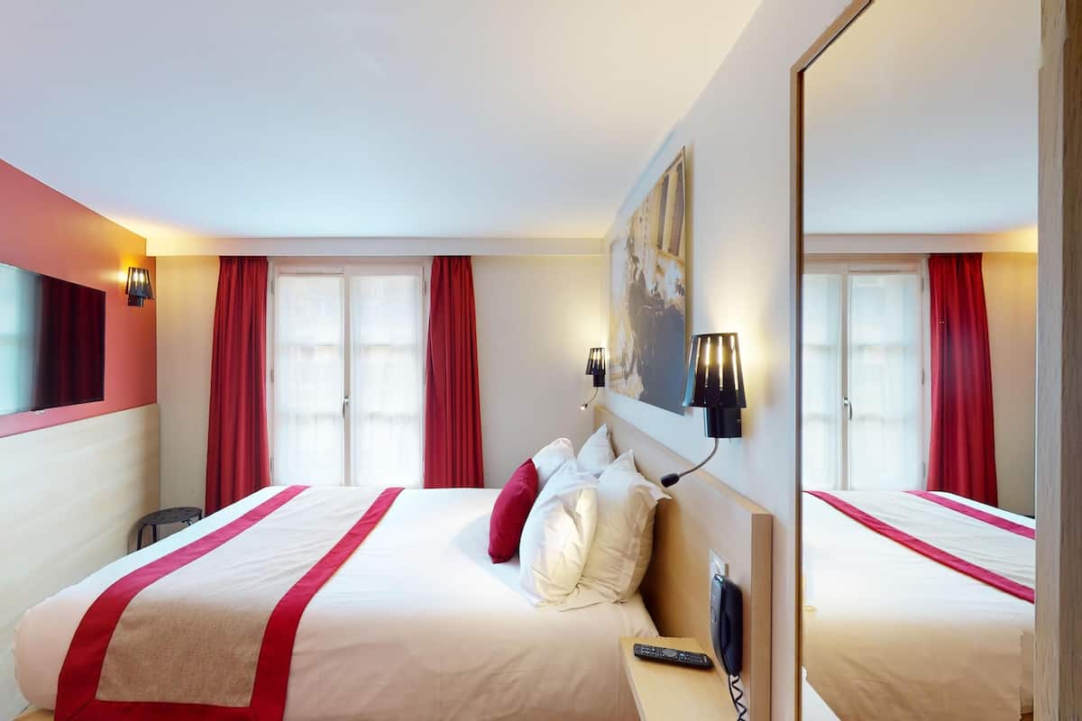 A hotel room with a large bed, a flat-screen TV mounted on the wall, a bedside table with a phone, and a window with red curtains.