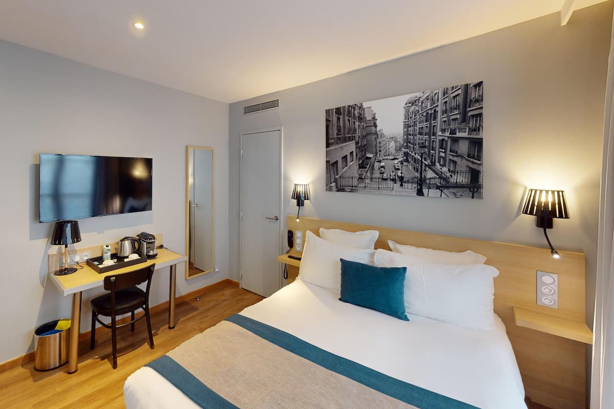A hotel room with a bed, a desk with a chair, a television, a coffee maker, and a cityscape painting on the wall.