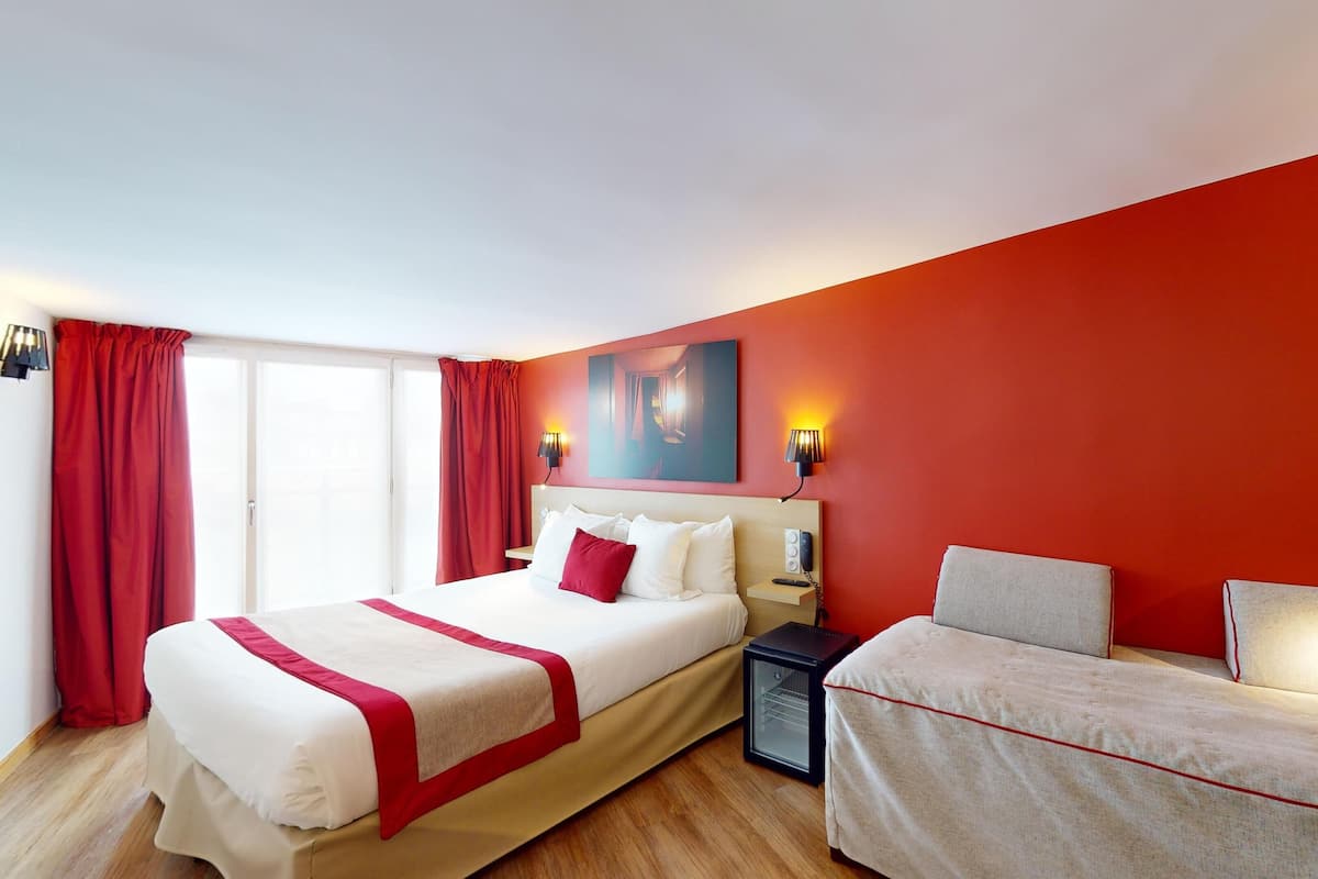 A hotel room with a large bed, a smaller bed, a bedside table with a lamp, a painting on the wall, and a window with red curtains.