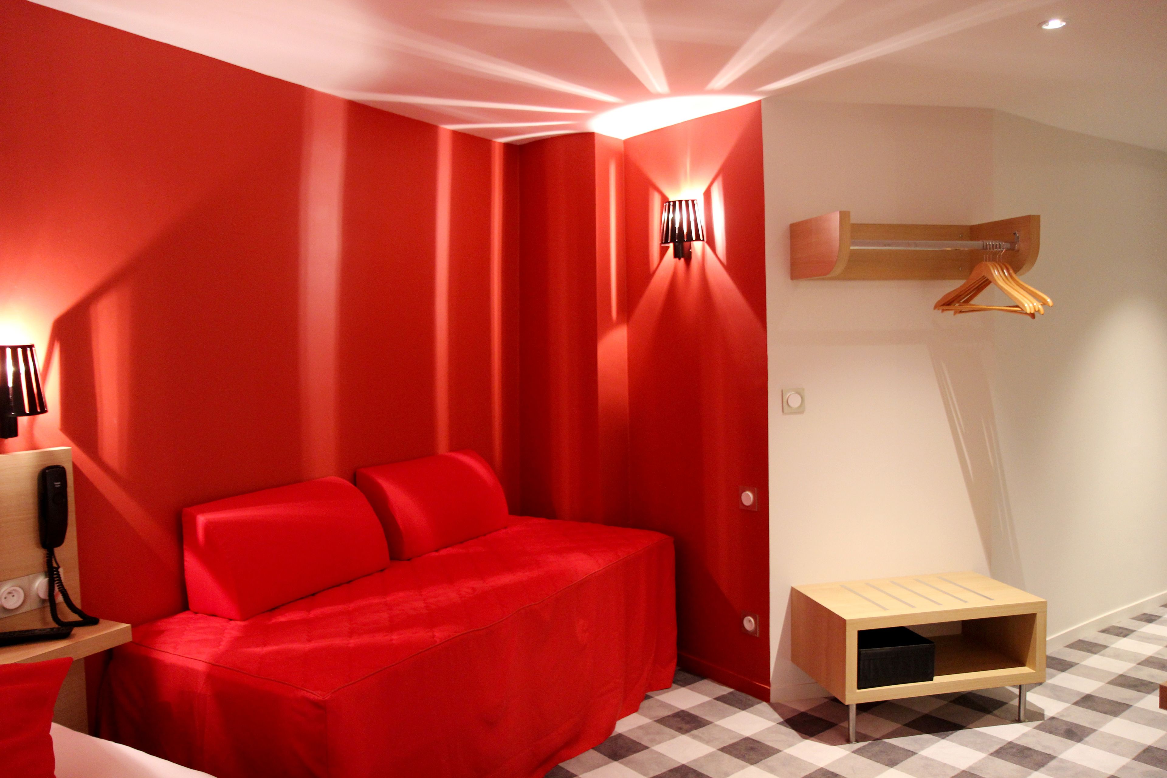 Habitación con decoración roja, una cama, una mesita de noche con lámpara y un mueble auxiliar de madera.