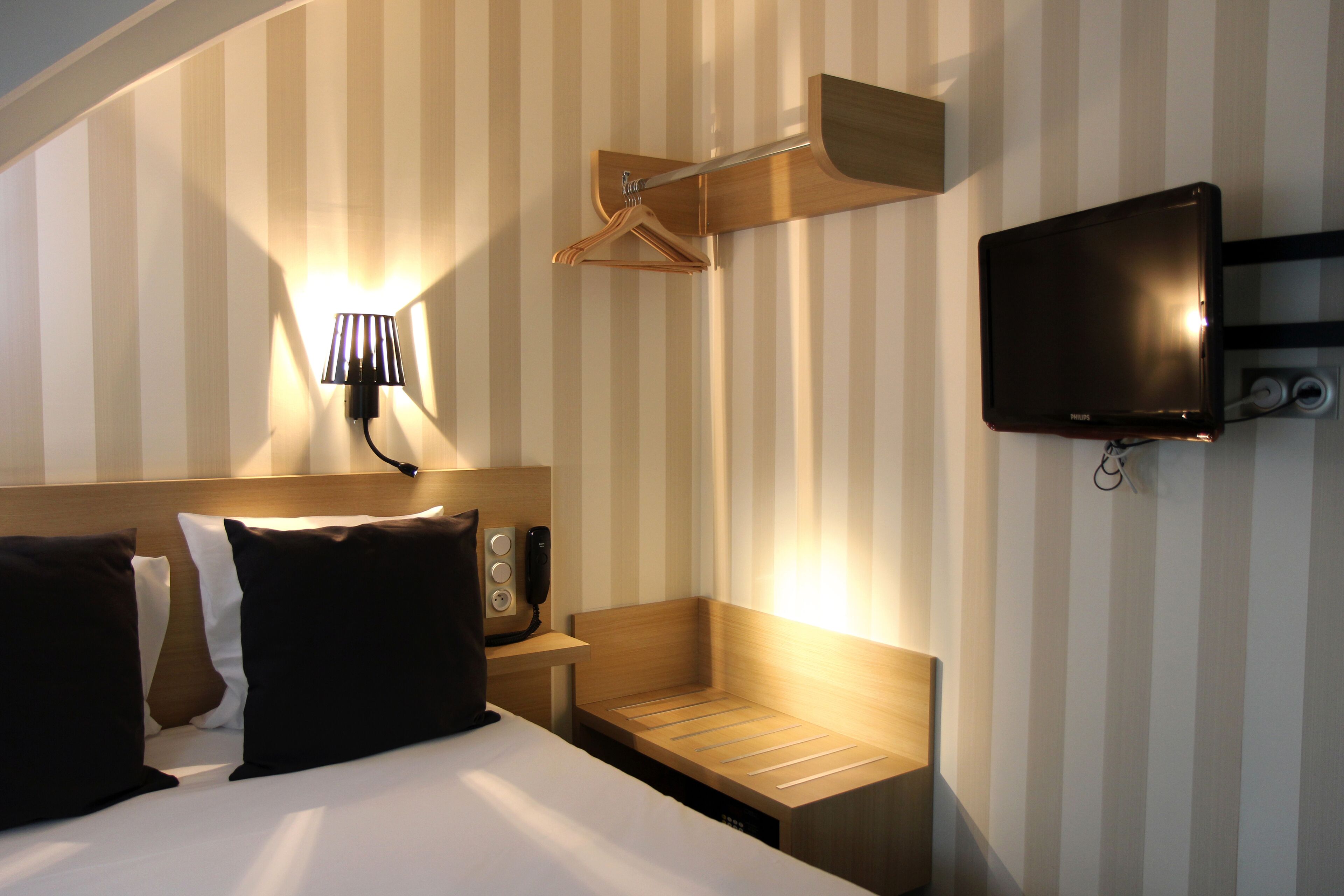A hotel room with a bed, bedside table, lamp, TV, and a wooden bench.