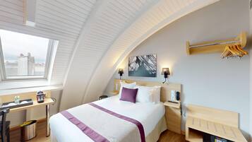 Ein modernes Hotelzimmer mit gewölbter Decke, einem großen Bett mit weißer und violetter Bettdecke, einem Schreibtisch mit Computer, einem Stuhl und einem Fenster mit Blick auf Gebäude.