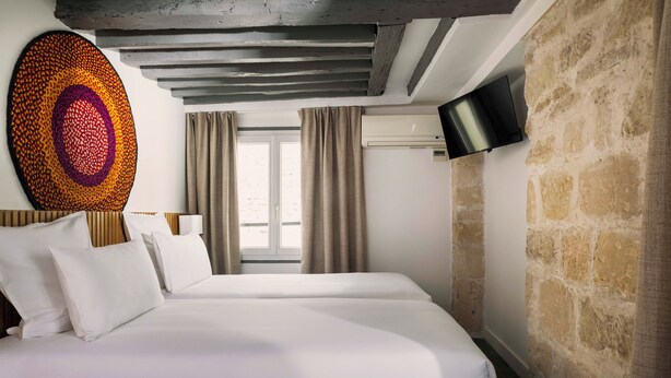A modern hotel room with a large bed, a flat-screen TV mounted on the wall, and a stone accent wall. - Korner Louvre (Paris)