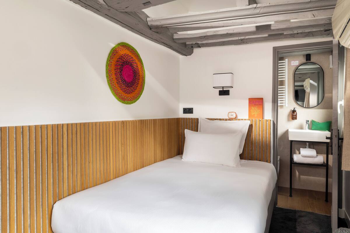 Hotelkamer met een houten hoofdbord, een bed met witte lakens, een kleurrijk kunstwerk aan de muur, een klein nachtkastje met een lamp, en een badkamer met spiegel en wastafel.