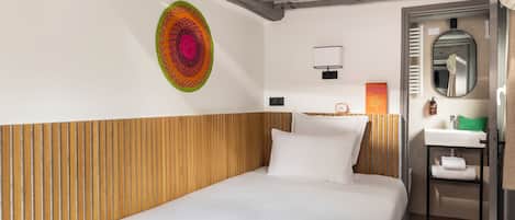 A hotel room with a wooden headboard, a bed with white linens, a colorful wall art piece, a small bedside table with a lamp, and a bathroom with a mirror and sink.