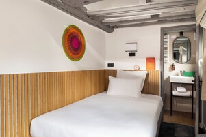 A hotel room with a wooden headboard, a bed with white linens, a colorful wall art piece, a small bedside table with a lamp, and a bathroom with a mirror and sink.
