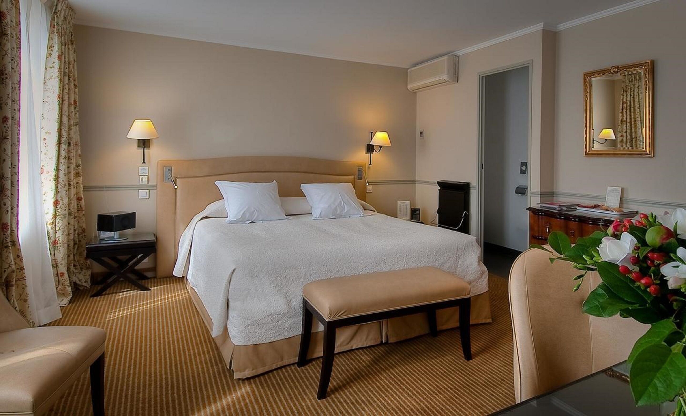 A hotel room with a large bed, two bedside lamps, a bench, a desk, a chair, a mirror, and a door.