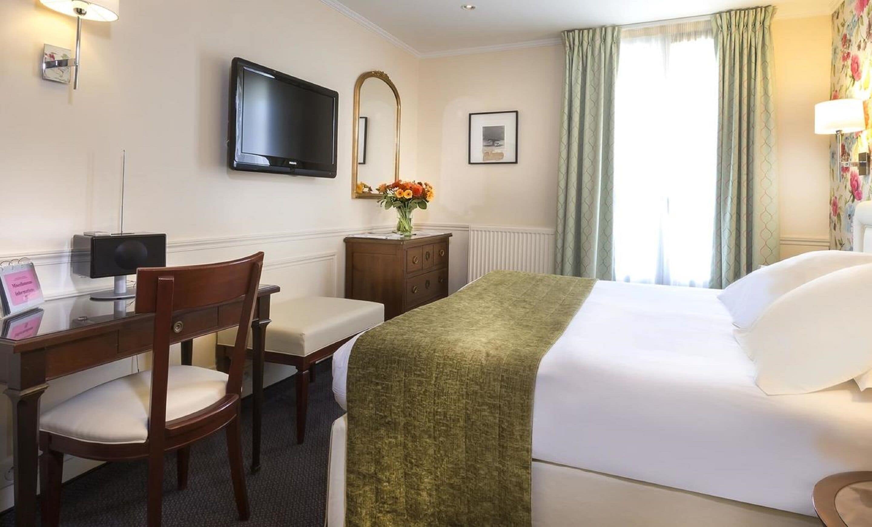 Ein Hotelzimmer mit einem Bett, einem Schreibtisch mit Stuhl, einem Fernseher, einem Spiegel und einem Fenster mit Vorhängen.