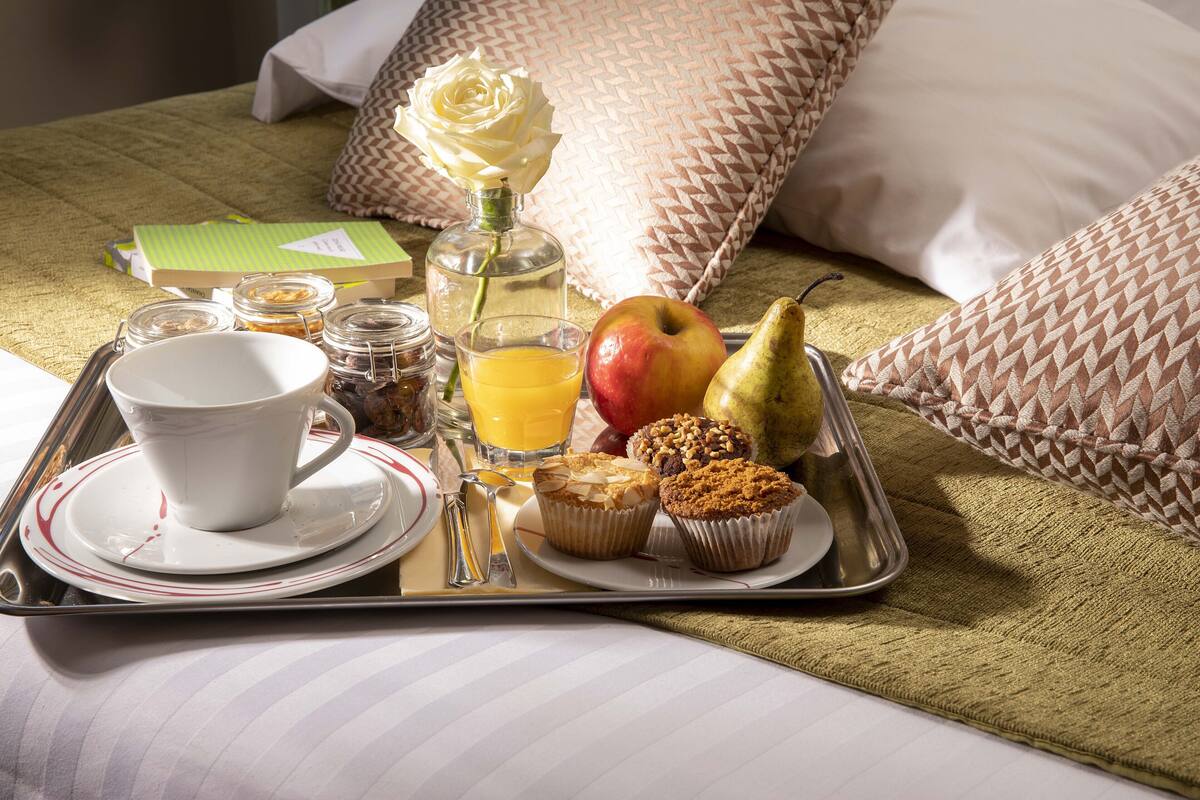 A breakfast tray with a cup, juice, apple, pear, muffins, and a bottle on a bed with patterned pillows.