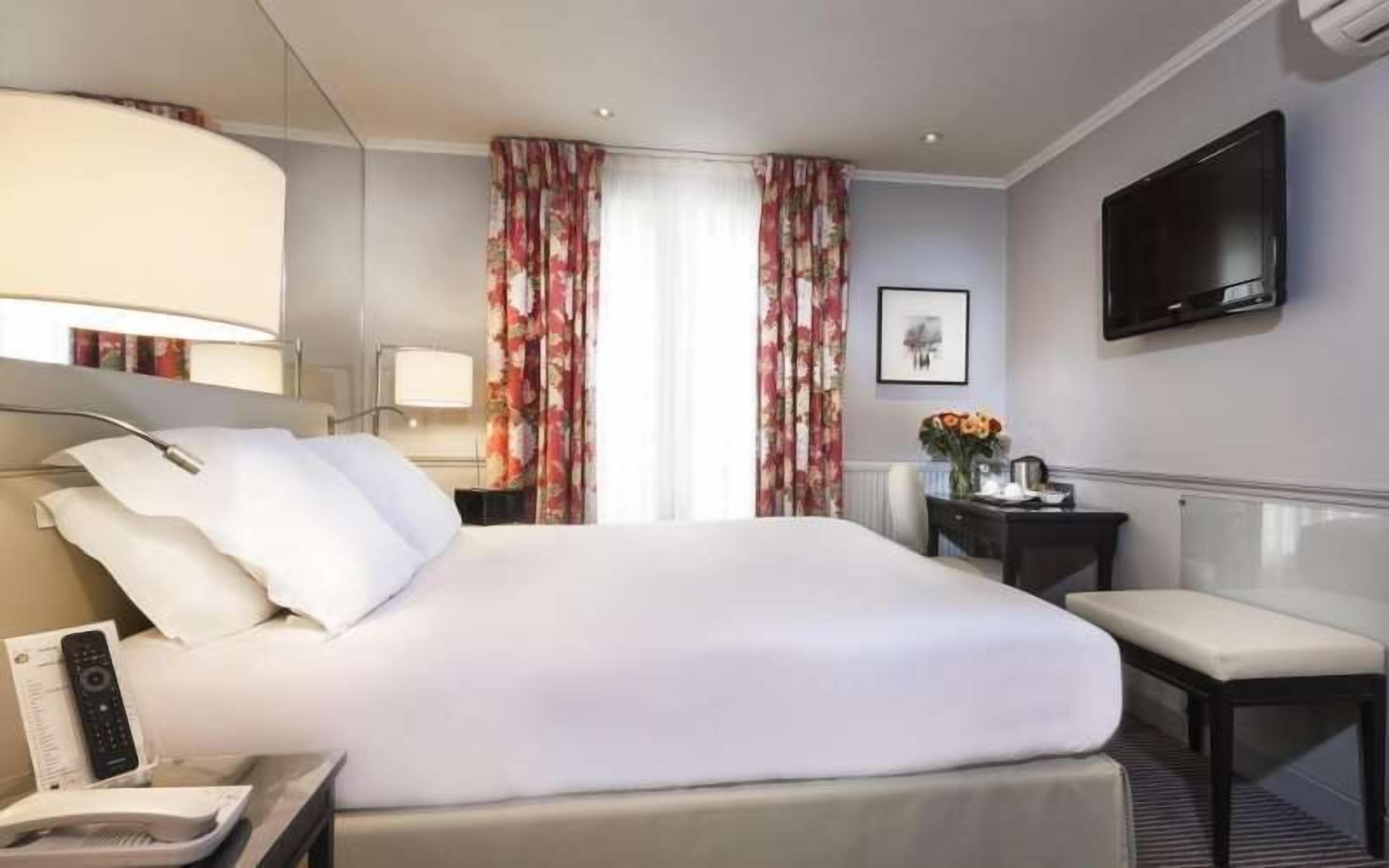 Quarto de hotel com uma cama grande, mesa de cabeceira com abajur, TV fixada na parede e janela com cortinas florais.