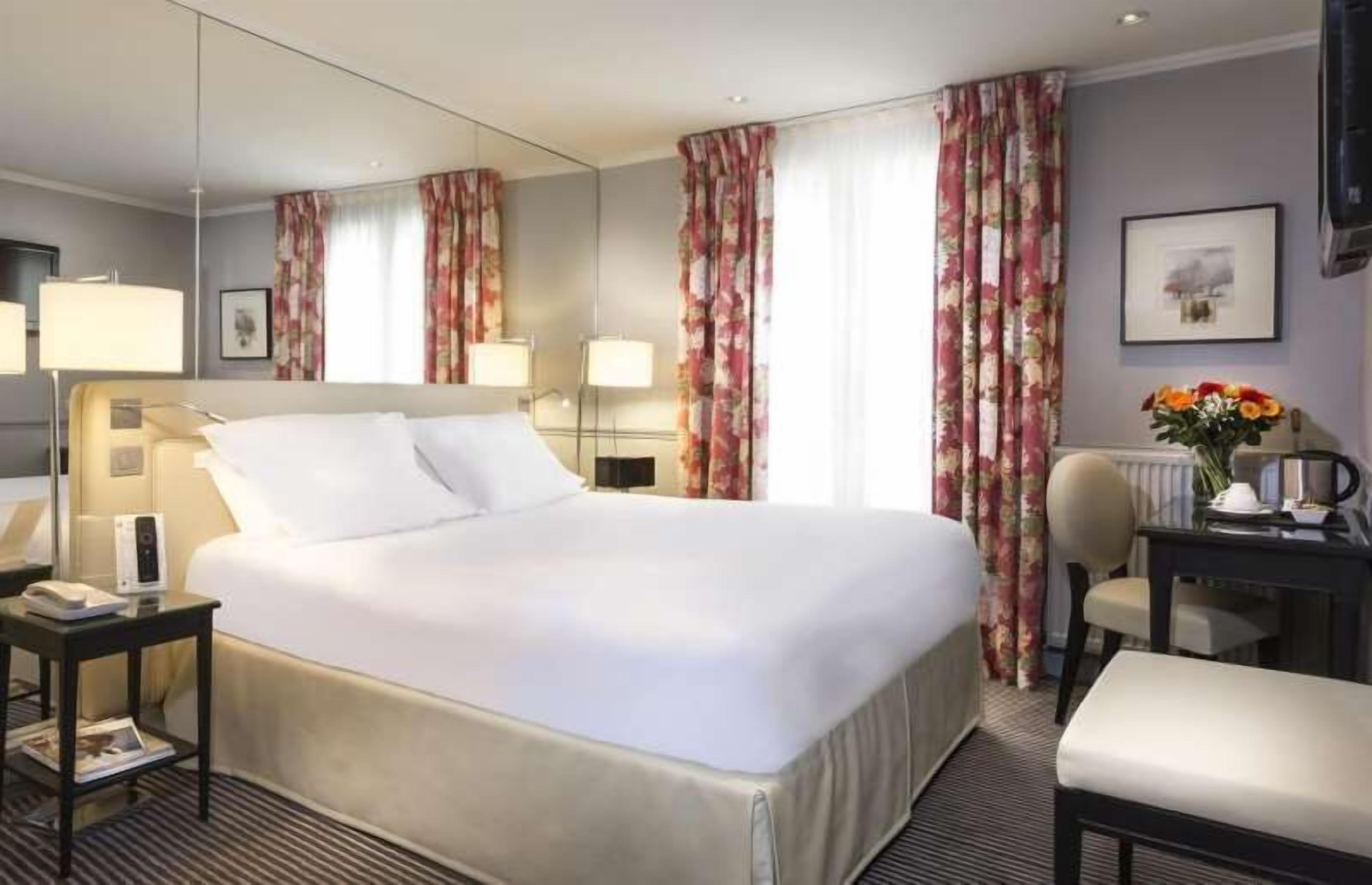 Habitación de hotel con una cama grande, una mesita de noche con lámpara, una silla, un escritorio con un jarrón de flores y un cuadro en la pared.