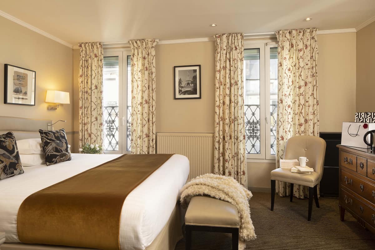 A hotel room with a large bed, a chair, a dresser, and two windows with floral curtains.