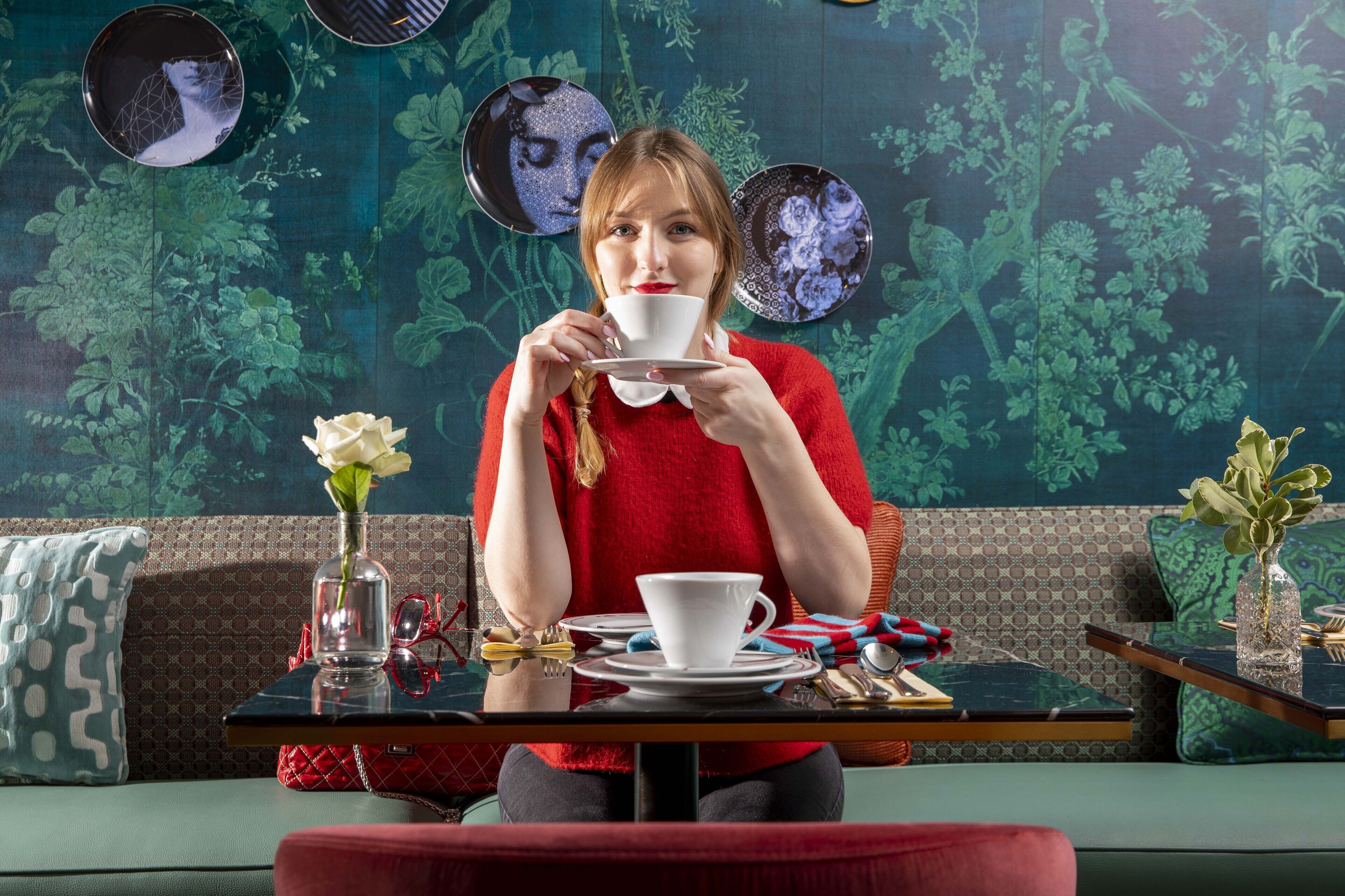 Une personne est assise à une table devant une tasse, un vase avec une fleur, et un mur à motifs en arrière-plan.